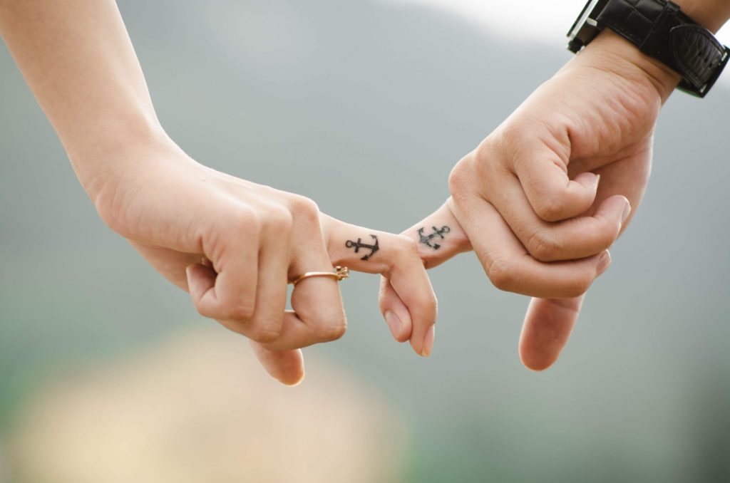 Two people with tattoos of anchors interlocking fingers. They are wearing a watch and a ring.