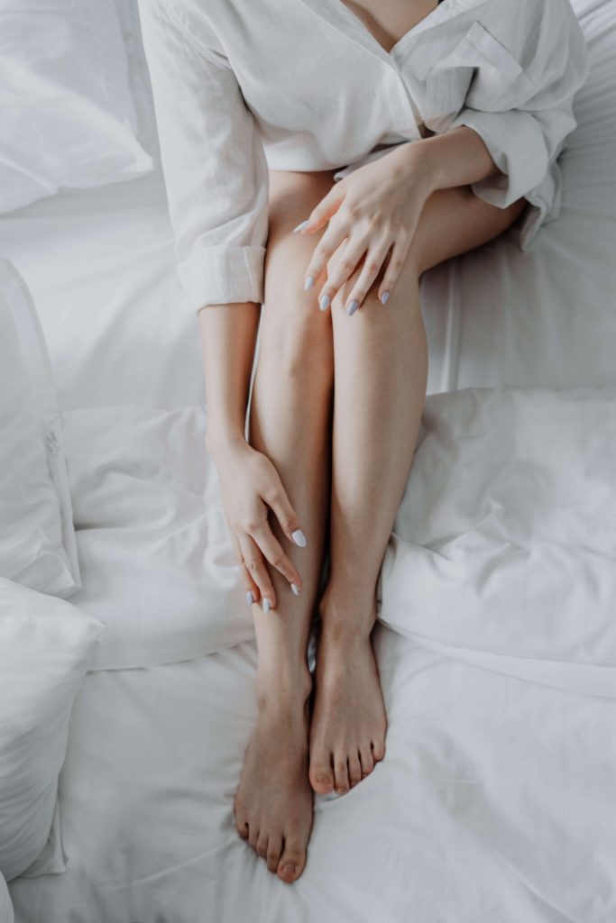 White woman with painted white nails sat holding her legs on the bed with a white shirt on